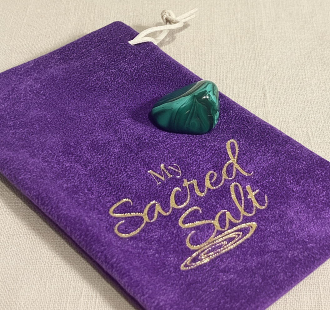 Malachite tumble stone on a purple pouch labeled 'My Sacred Salt' on a cream linen background